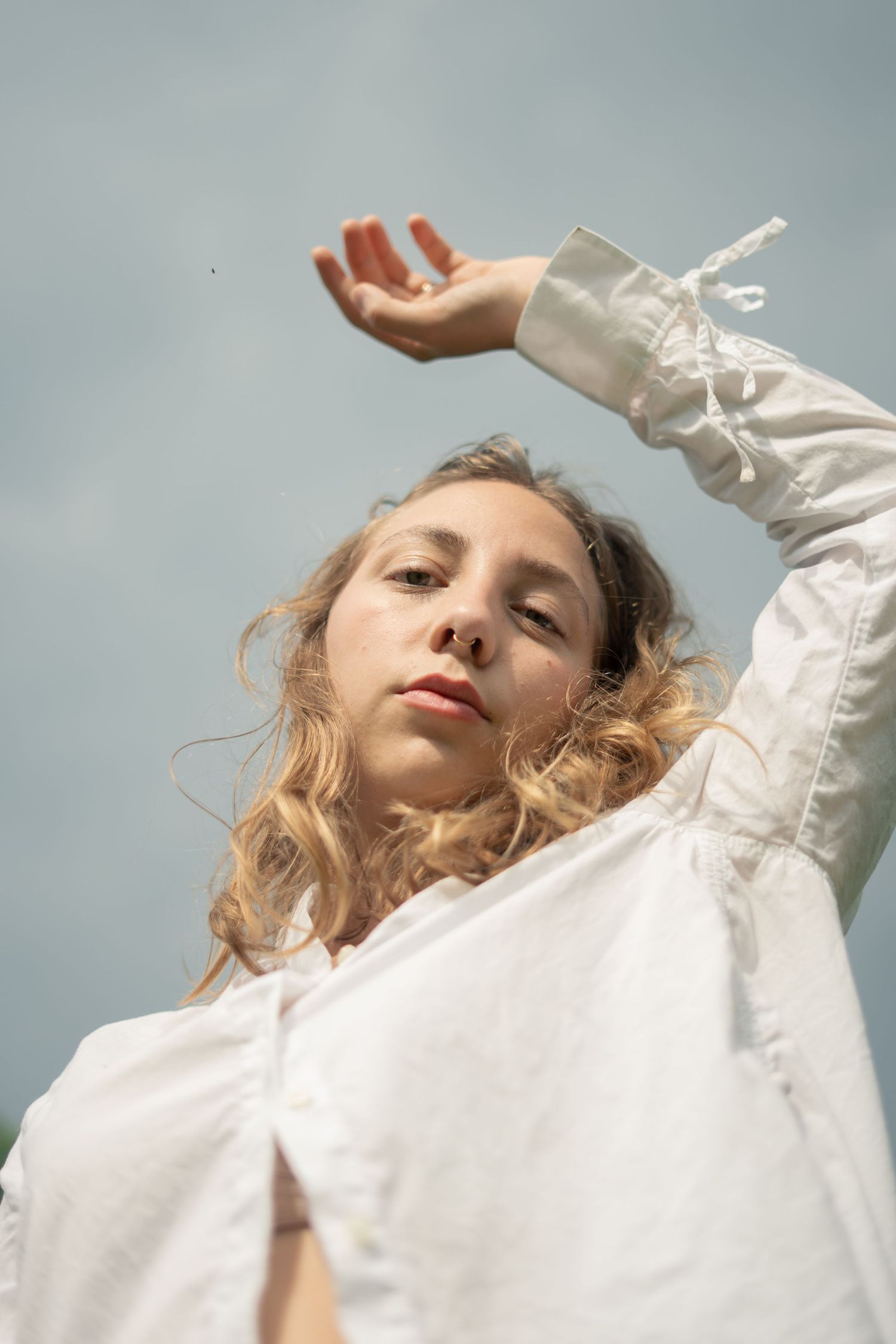 Low angle portrait of young woman in white ruffled shirt with arm raised against cloudy sky background.