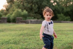 A toddler in striped shirt and bow tie walks across grassy field.