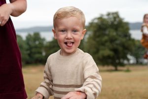 A young child wearing a beige sweater smiles joyfully while playing outdoors on a grassy field.