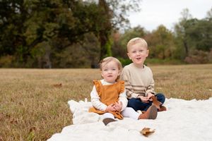 Two children sit on a white blanket in a grassy field during fall.