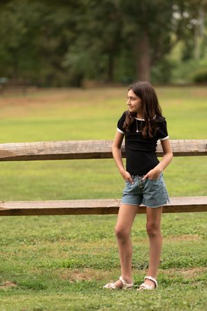 A person in denim shorts and black shirt leans against a wooden fence in a grassy field on a sunny day.