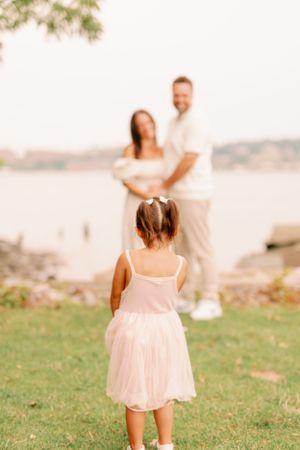 Child in pink dress facing couple by waterfront at sunset.