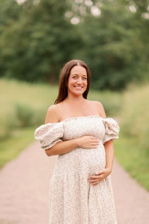 Expectant mother in white off-shoulder dress standing on rural path.