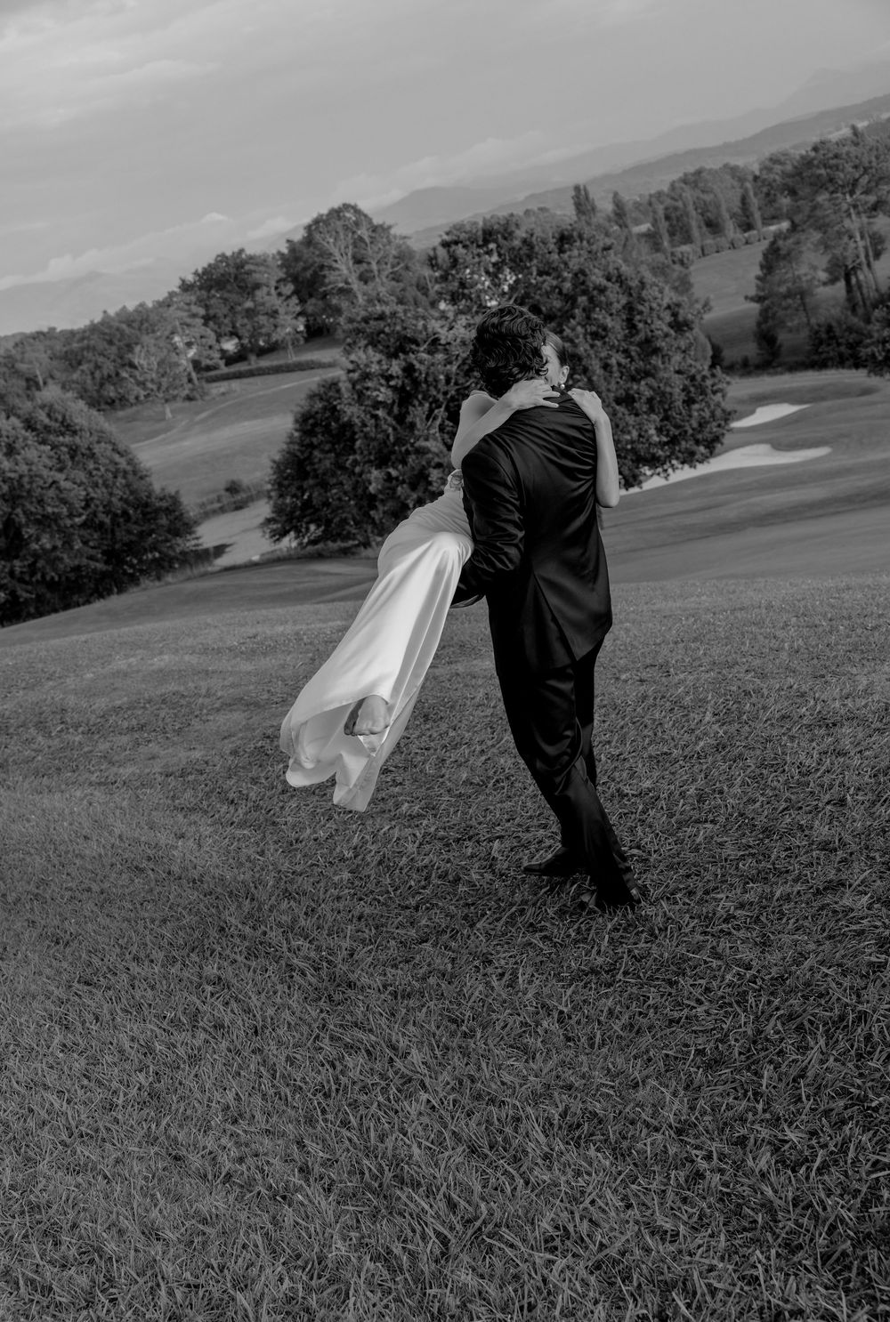 A couple embraces on a hillside with flowing white dress fabric caught in the wind, shot in black and white.