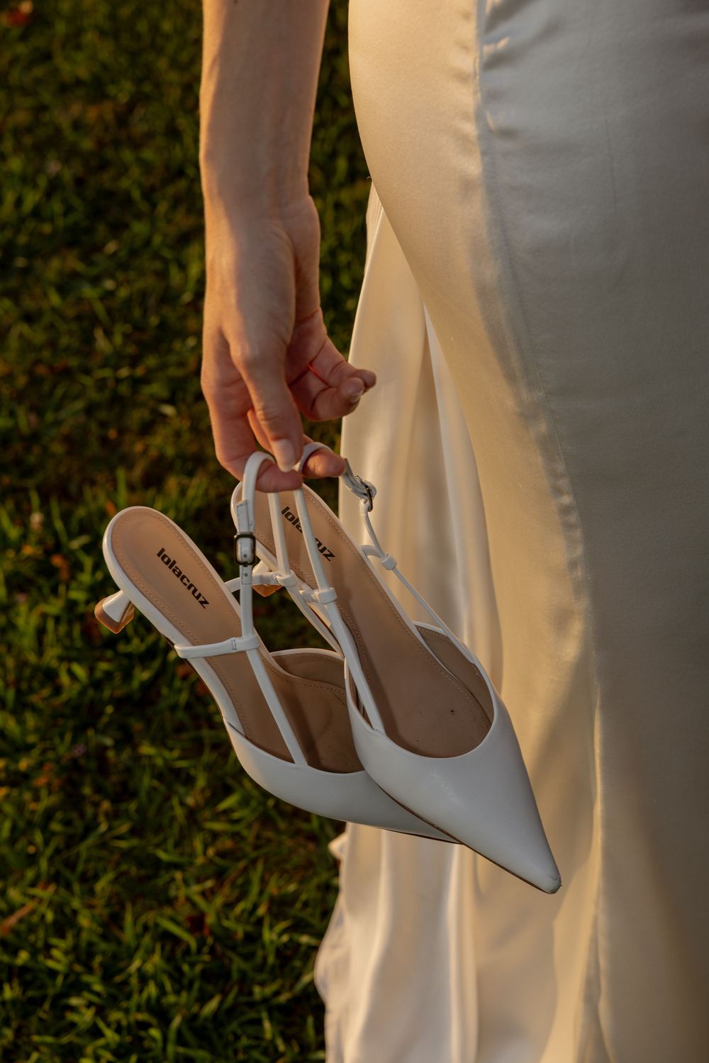 Close-up of a hand holding white pointed-toe kitten heel shoes against a white wedding dress.