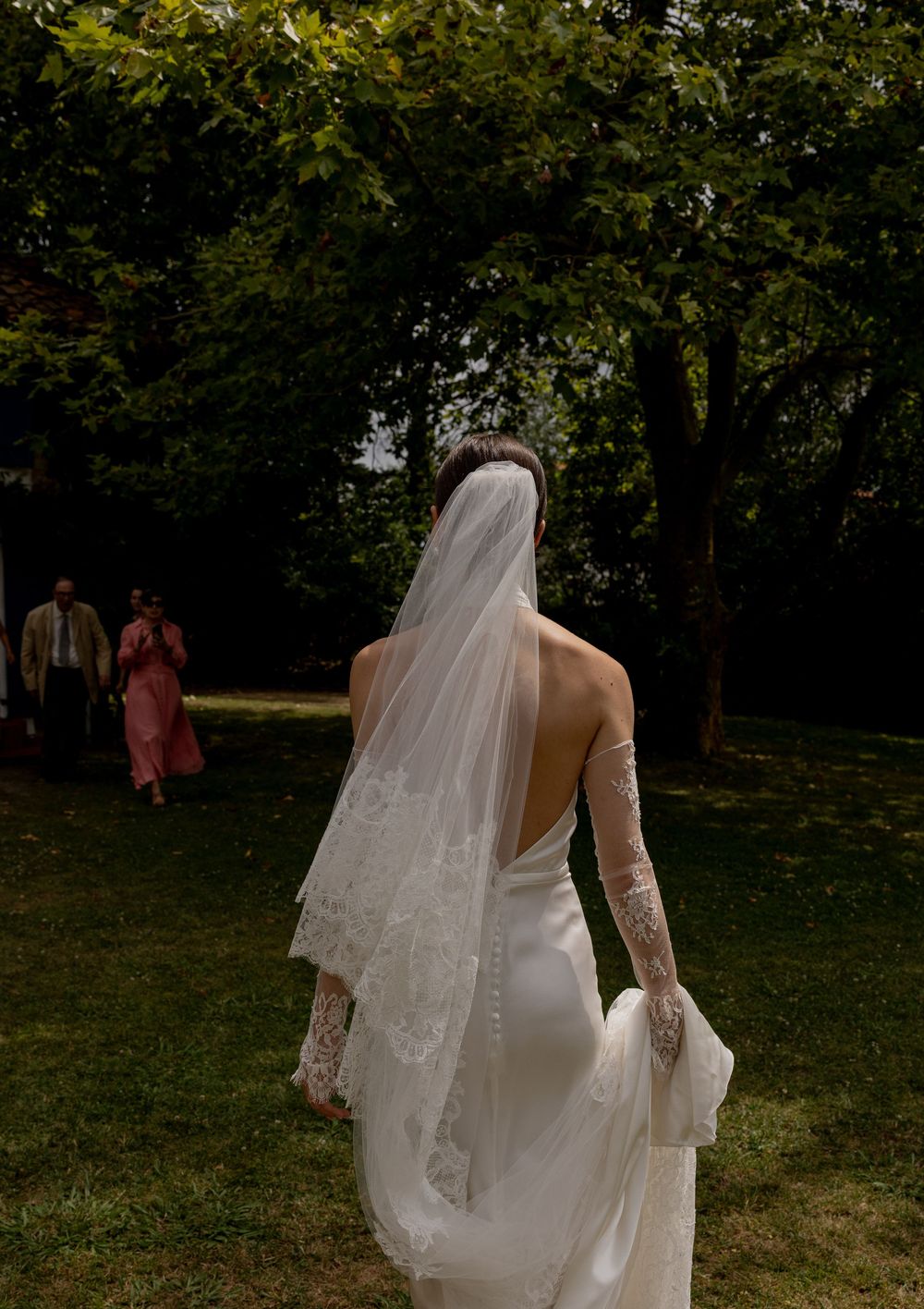 A bride in an elegant white dress with a flowing veil walks on grass while guests stand in the background.