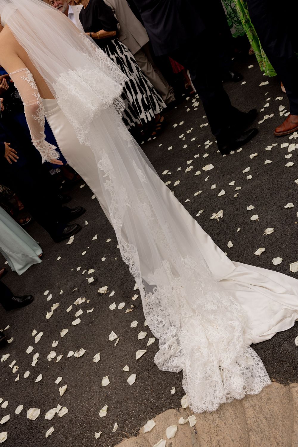 A flowing white lace wedding dress train trails on the ground surrounded by scattered flower petals.
