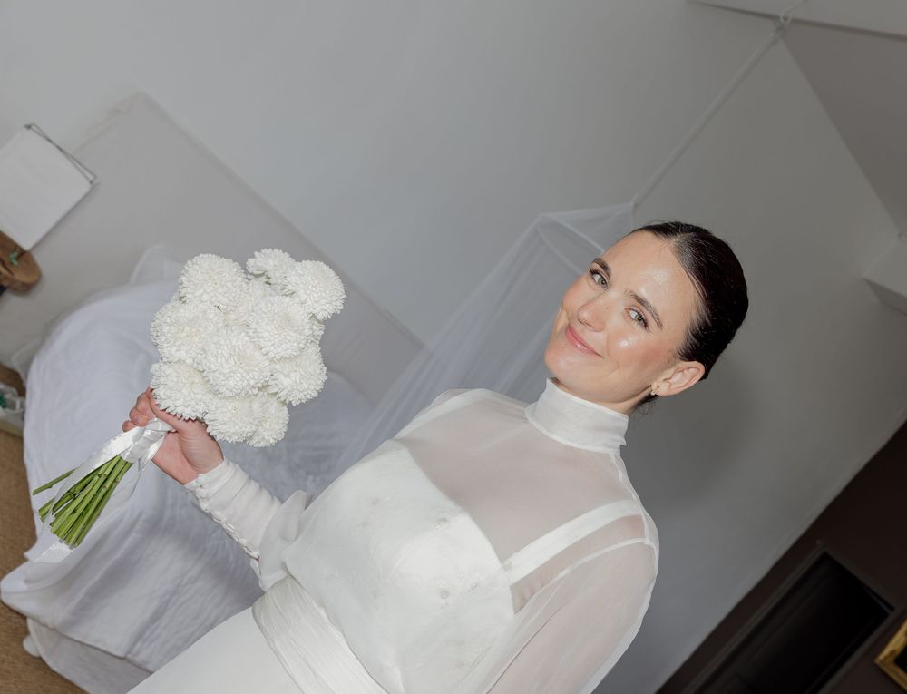 Bride in white sheer gown holding ivory bouquet with veil, elegant updo hairstyle, posing indoors.