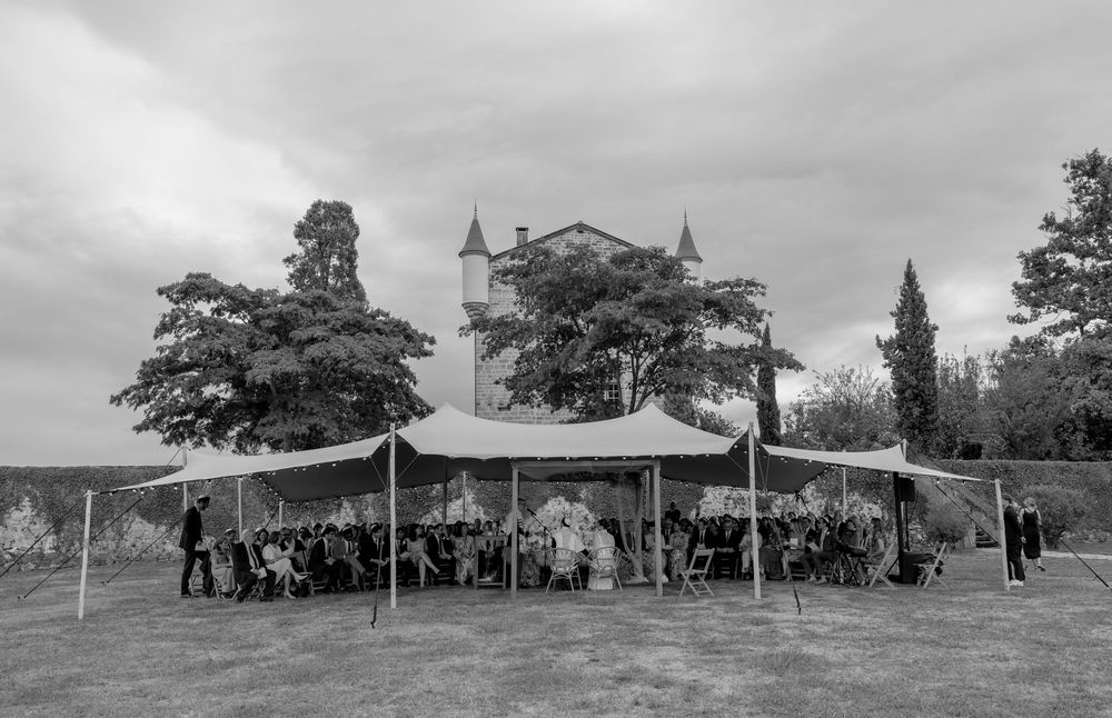 Black and white photo of an outdoor wedding reception under a large tent near a historic château surrounded by trees.