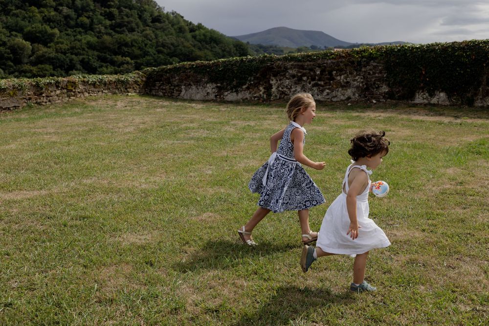 Two young girls in dresses running on green grass near stone ruins with hills in background.