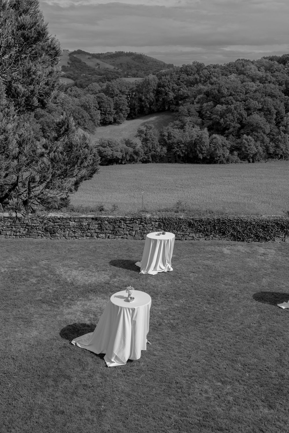 Black and white photo of two draped cocktail tables on a lawn with rolling hills and lush trees in the background.
