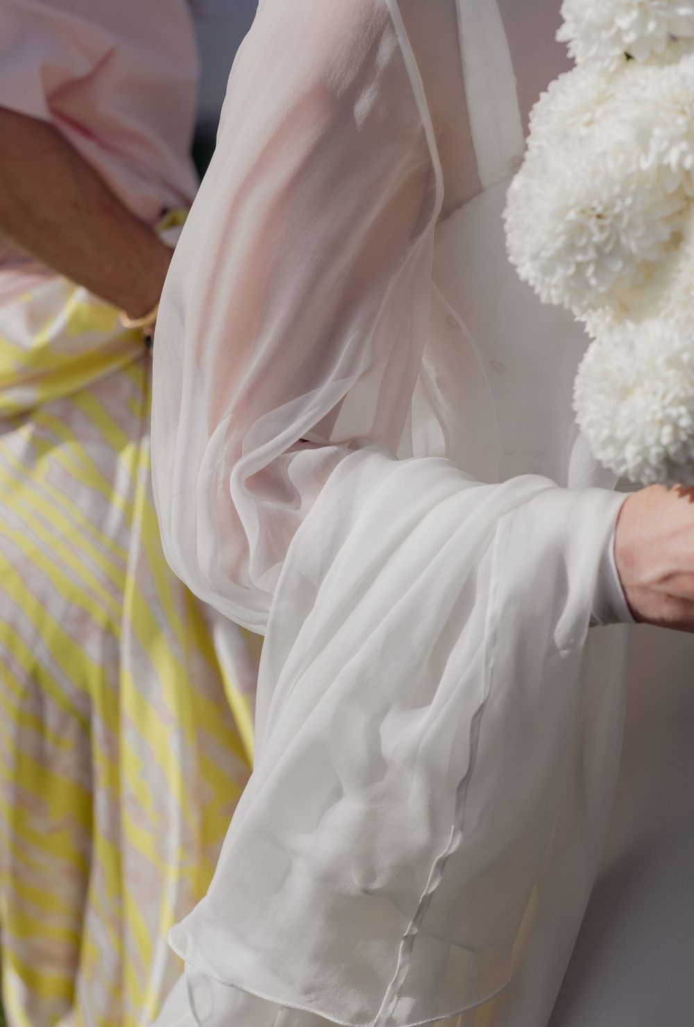 Bride in flowing white chiffon gown holding white floral bouquet beside guest in yellow striped dress.