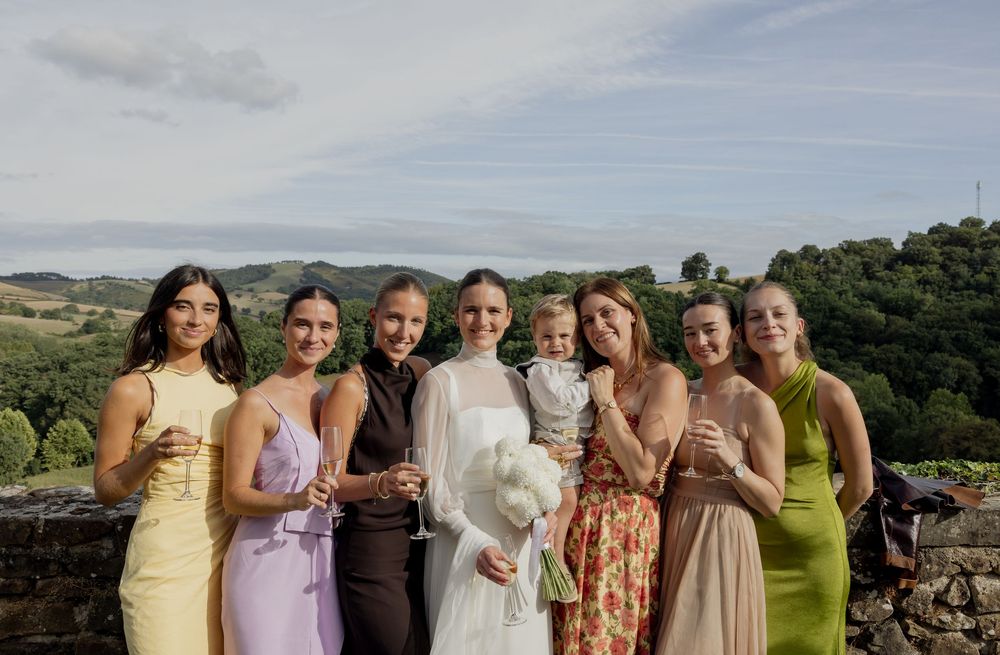 Bride in white gown poses with bridesmaids in colorful dresses outdoors with scenic hills in background.