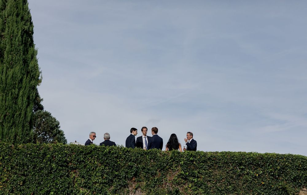 Group of people in formal attire conversing outdoors near lush green hedges and cypress trees under a cloudy sky.