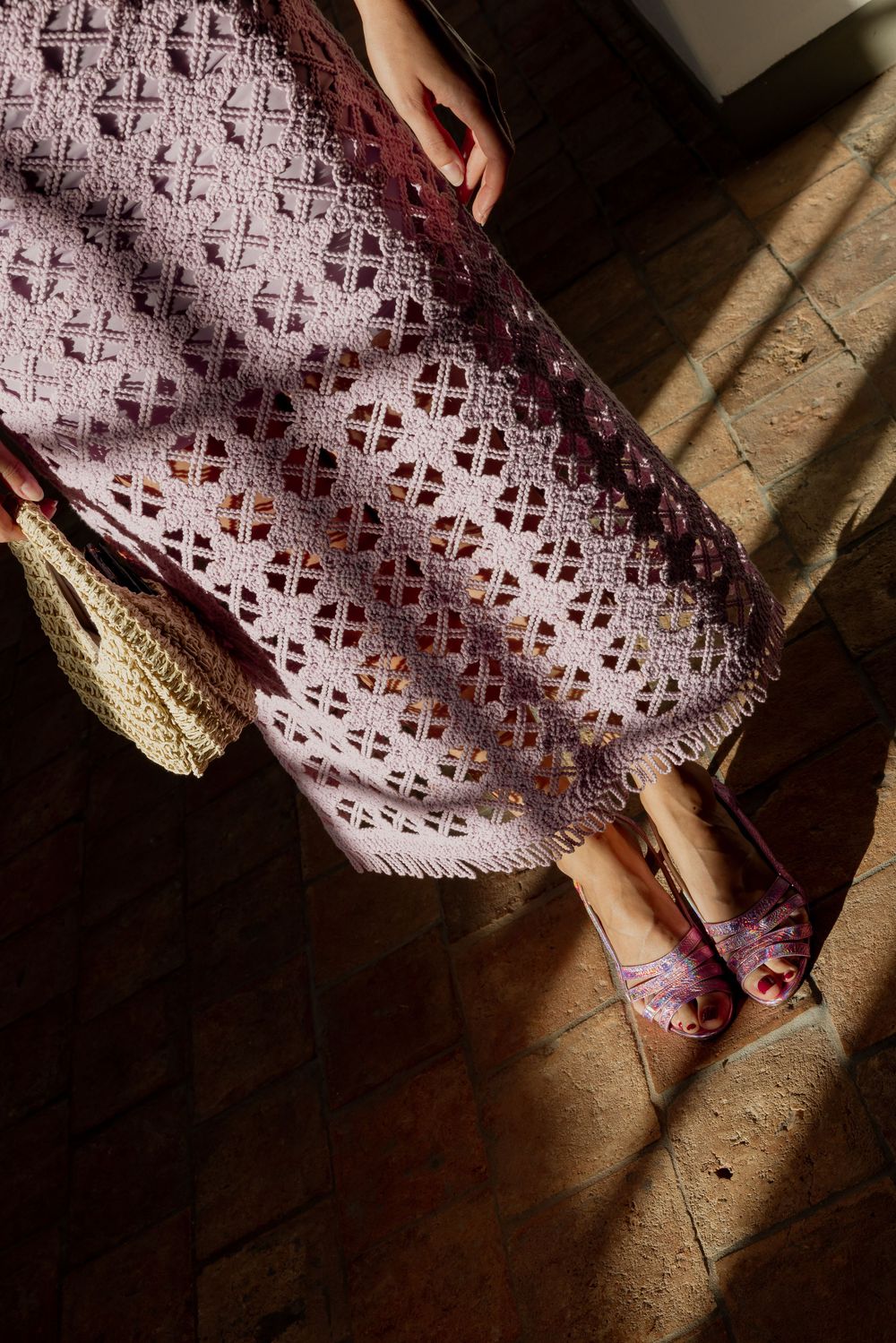 Woman wearing a pink crochet midi skirt with fringe hem, paired with purple heeled sandals and a woven bag.