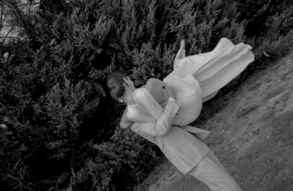 Black and white photo of a woman in a flowing white dress lying among evergreen shrubs outdoors.