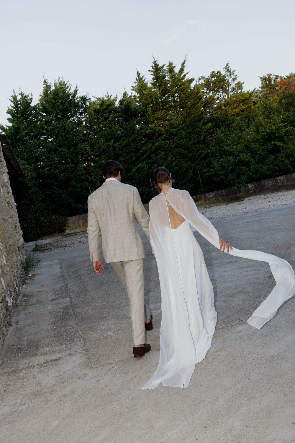 Bride and groom walking away in elegant white wedding dress with flowing cape and beige suit, surrounded by lush green trees.