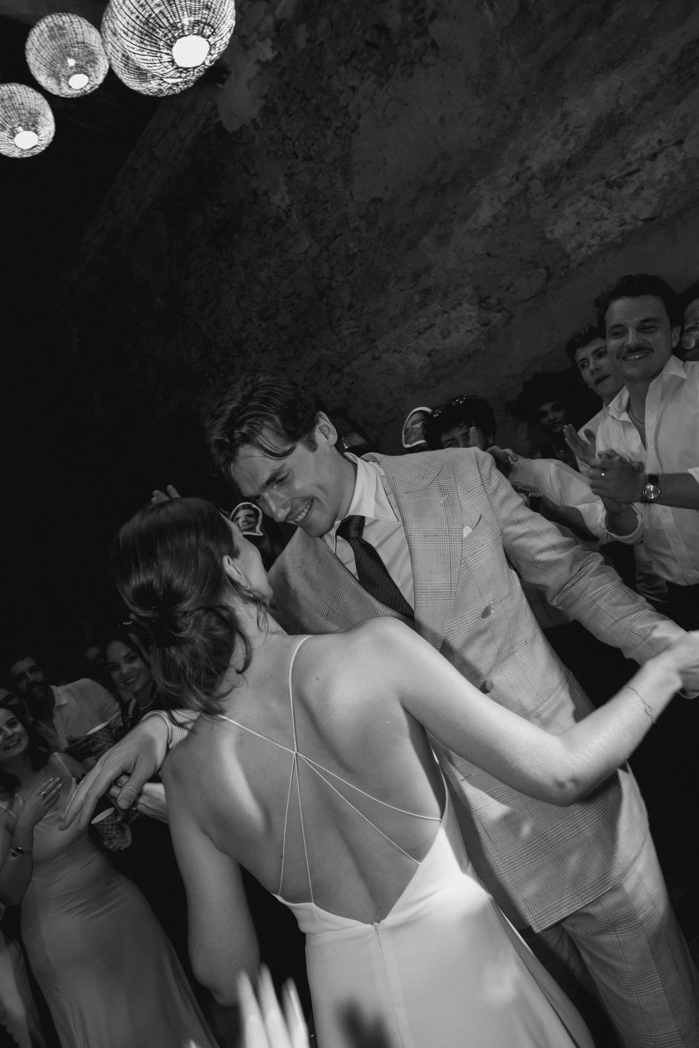 Black and white photo of a bride and groom sharing a joyful first dance surrounded by guests at their wedding reception.