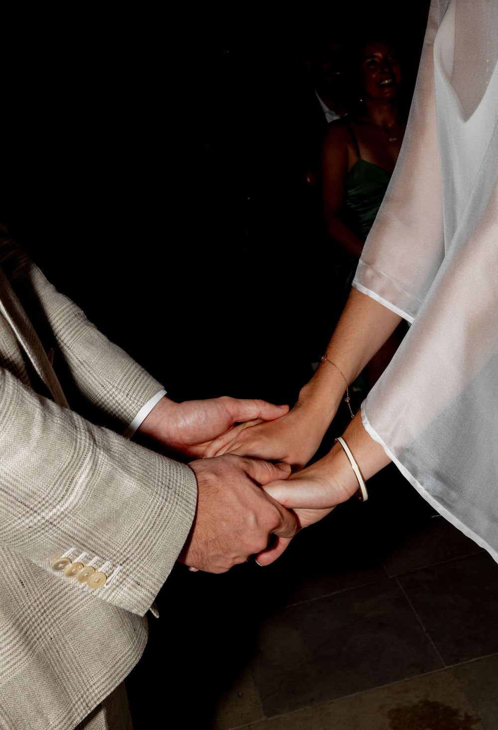 A couple holds hands tenderly at a wedding, the groom in a plaid suit and bride in a sheer white dress with a gold bracelet.