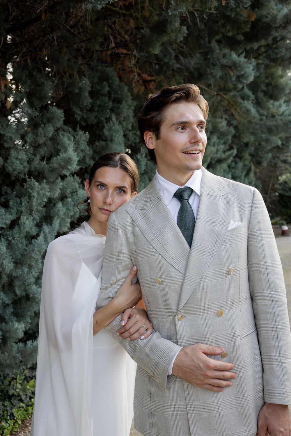 Elegant bride in white cape dress holds groom in light gray double-breasted suit with green tie outdoors.