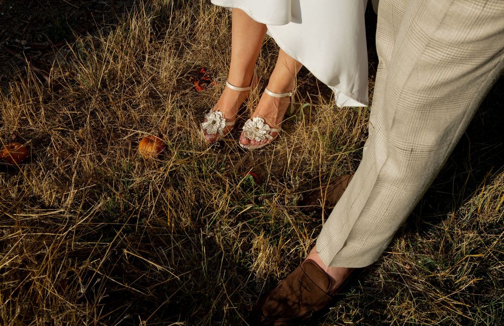 Close-up of bride in floral heels and groom in tan suit standing on dry grass at outdoor wedding.