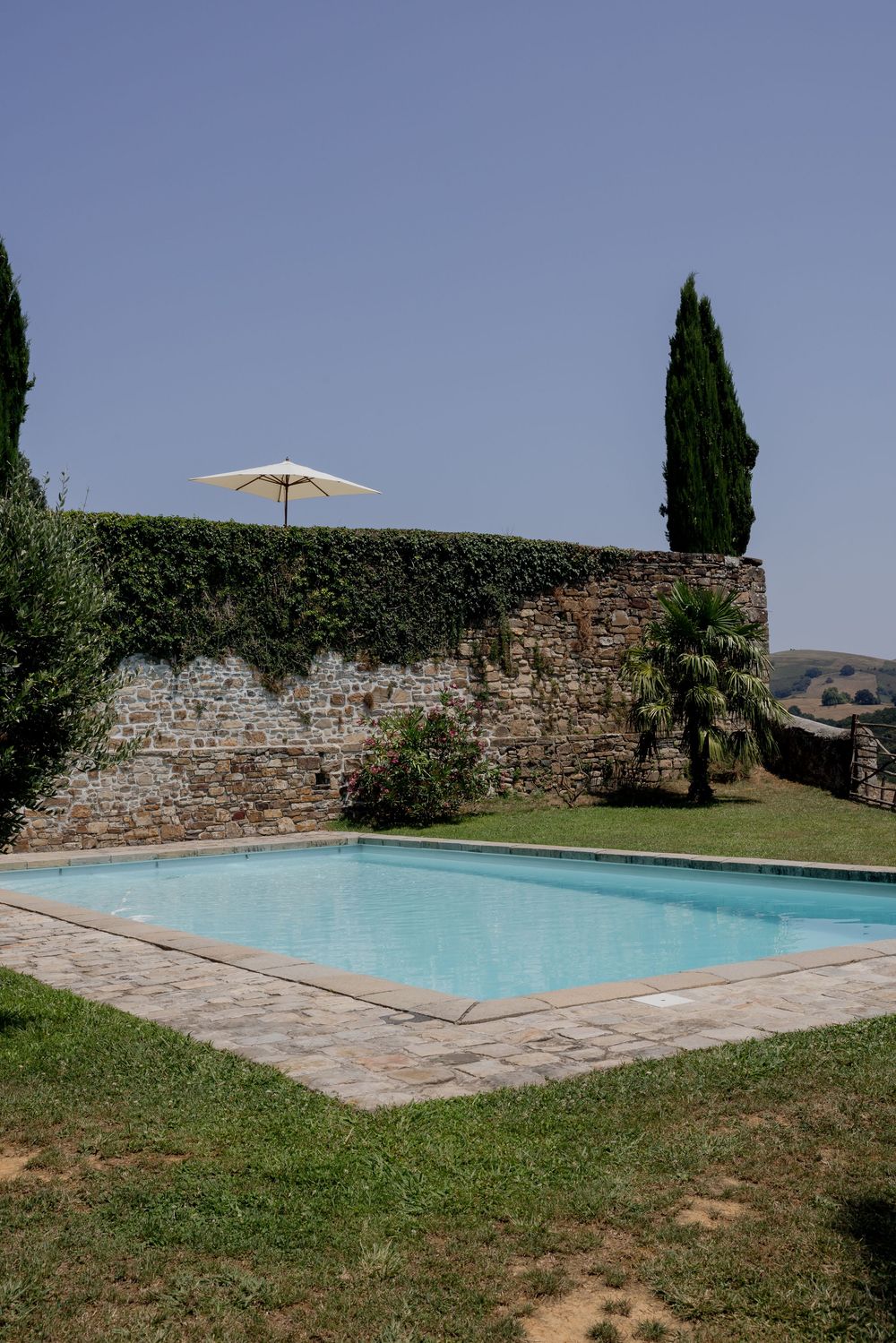 A rectangular swimming pool beside an ivy-covered stone wall with a white umbrella and cypress trees under clear blue sky.