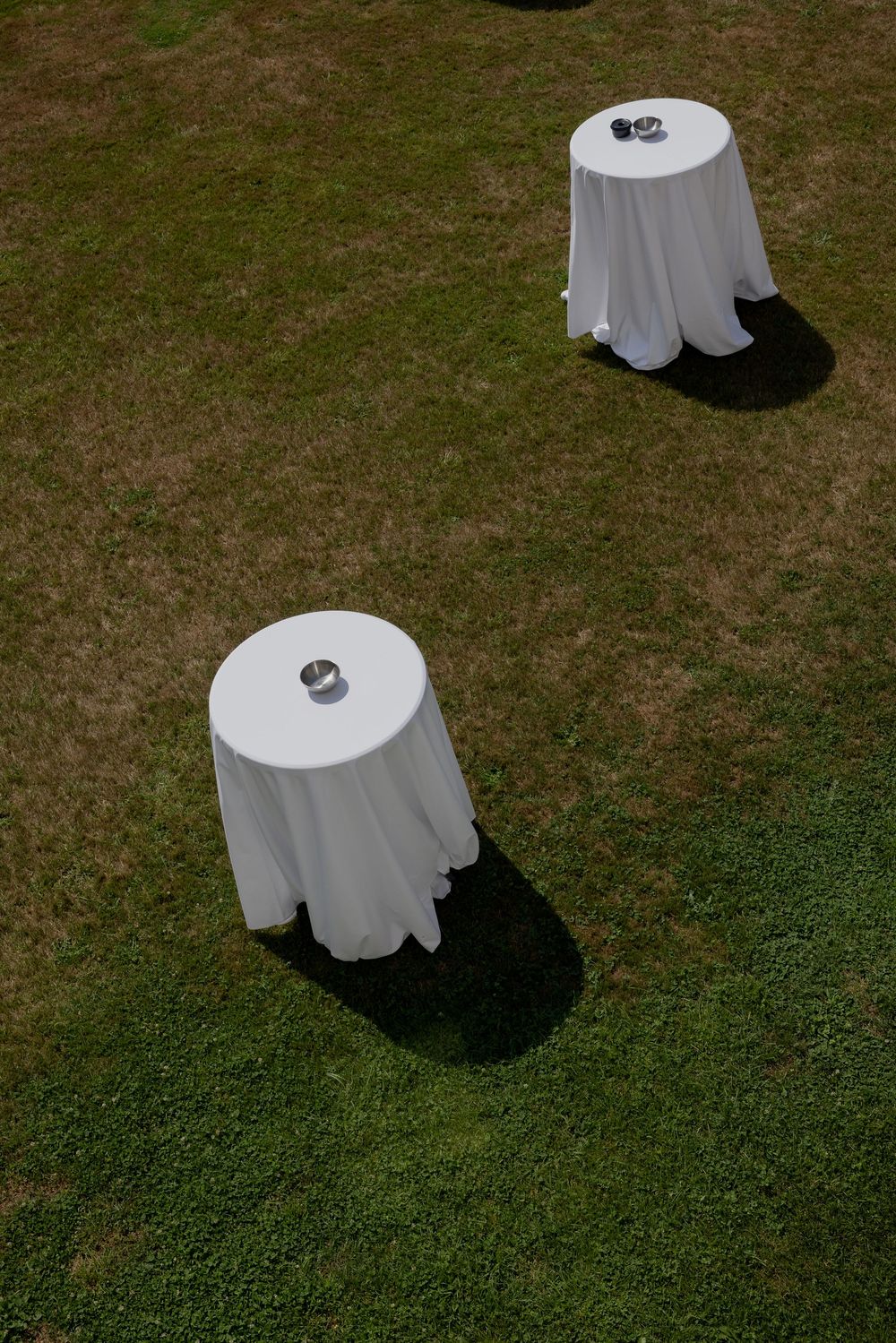 Two round cocktail tables with white tablecloths and small centerpieces set on green grass outdoors.