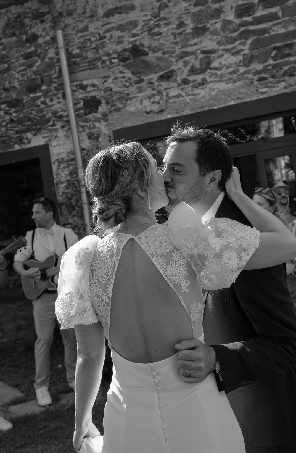 Black and white photo of a bride and groom sharing a kiss at their outdoor wedding near a stone building.