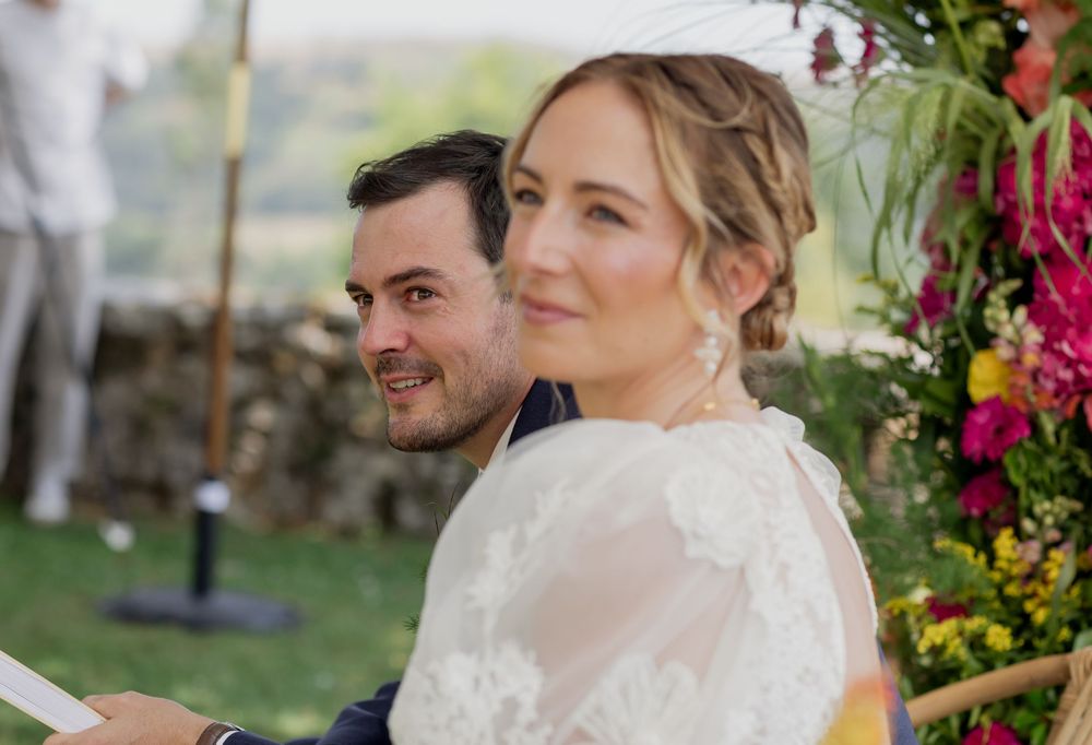 Bride and groom smiling at outdoor wedding ceremony with colorful floral backdrop.
