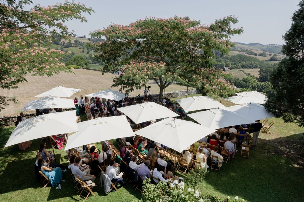 Outdoor wedding reception with white umbrellas, guests dining on a sunny hillside with scenic countryside views.