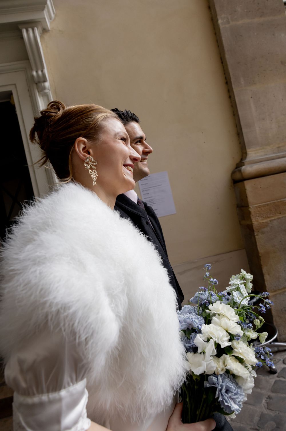 Smiling bride in white fur shawl holding blue and white bouquet stands with groom in black suit at wedding.