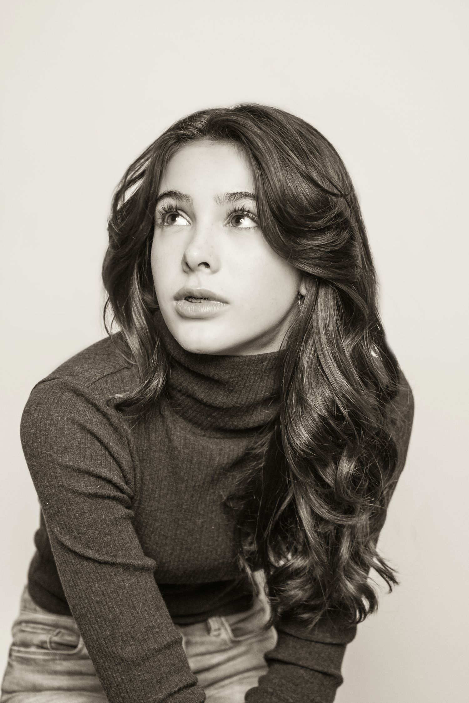 Portrait of a woman in brown turtleneck sweater looking up with wavy dark hair against light background.