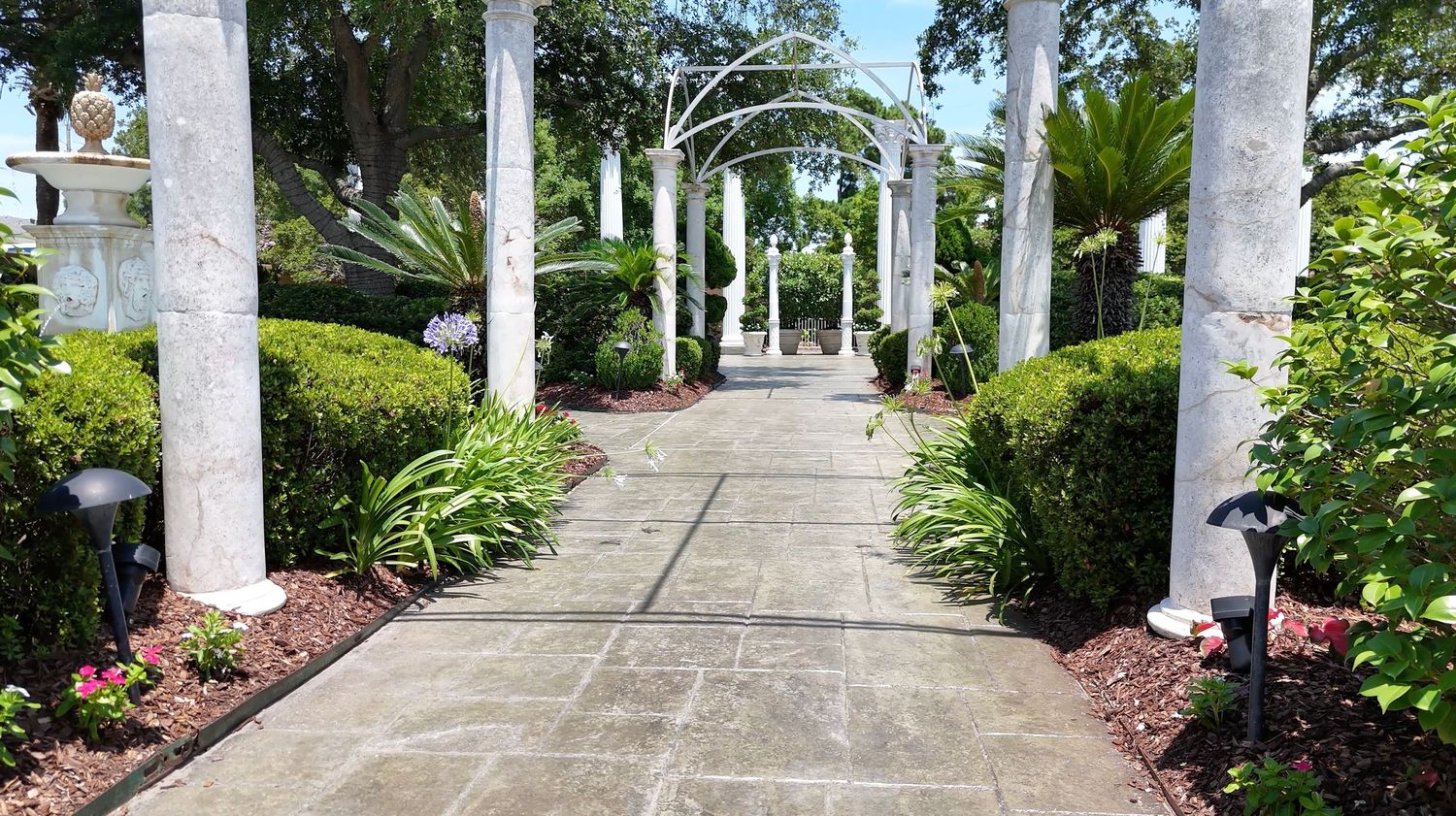 A picturesque garden walkway with white columns and pergolas leading to a gazebo.