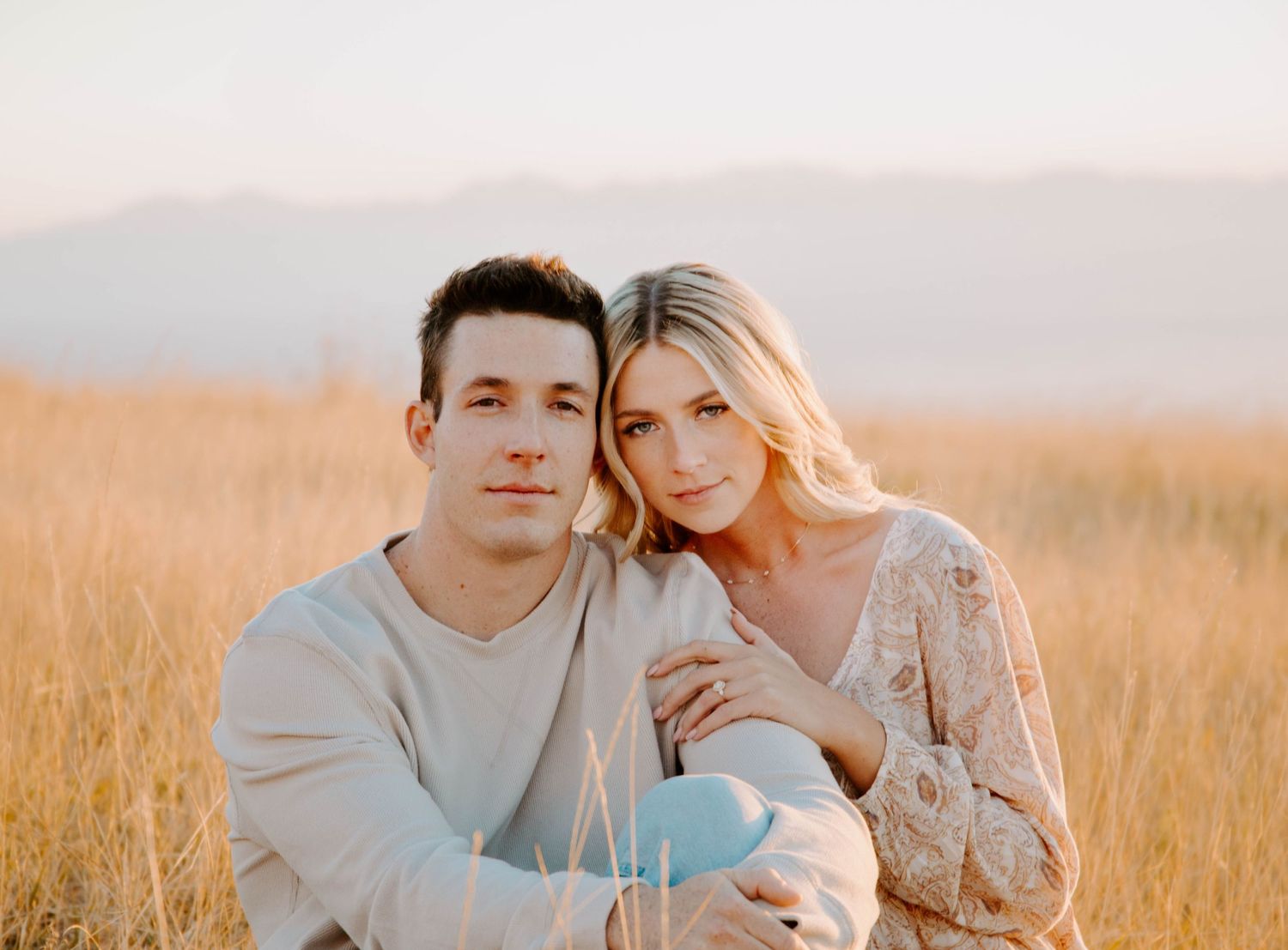 Romantic couple embraces in a golden wheat field at sunset wearing neutral tones.