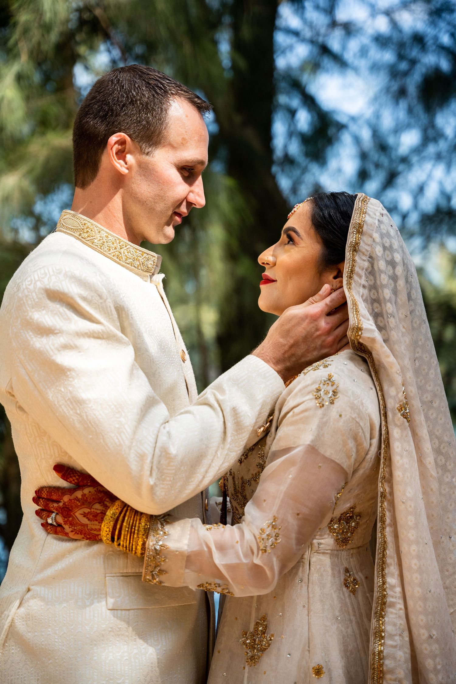 Intimate moment between couple in white and gold traditional wedding attire in forest setting.