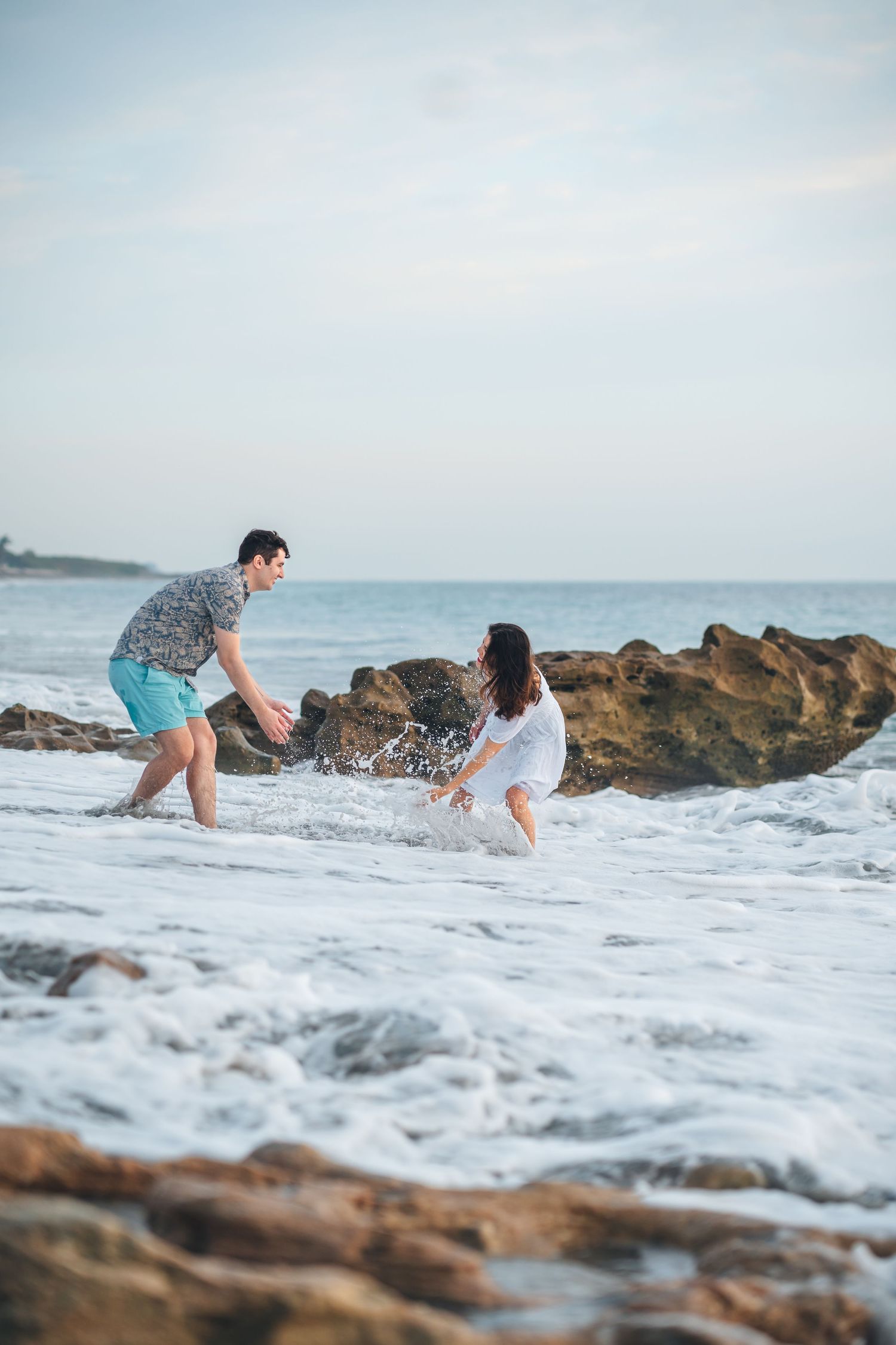 Playful couple splashing in ocean waves among rocky beach at sunset.