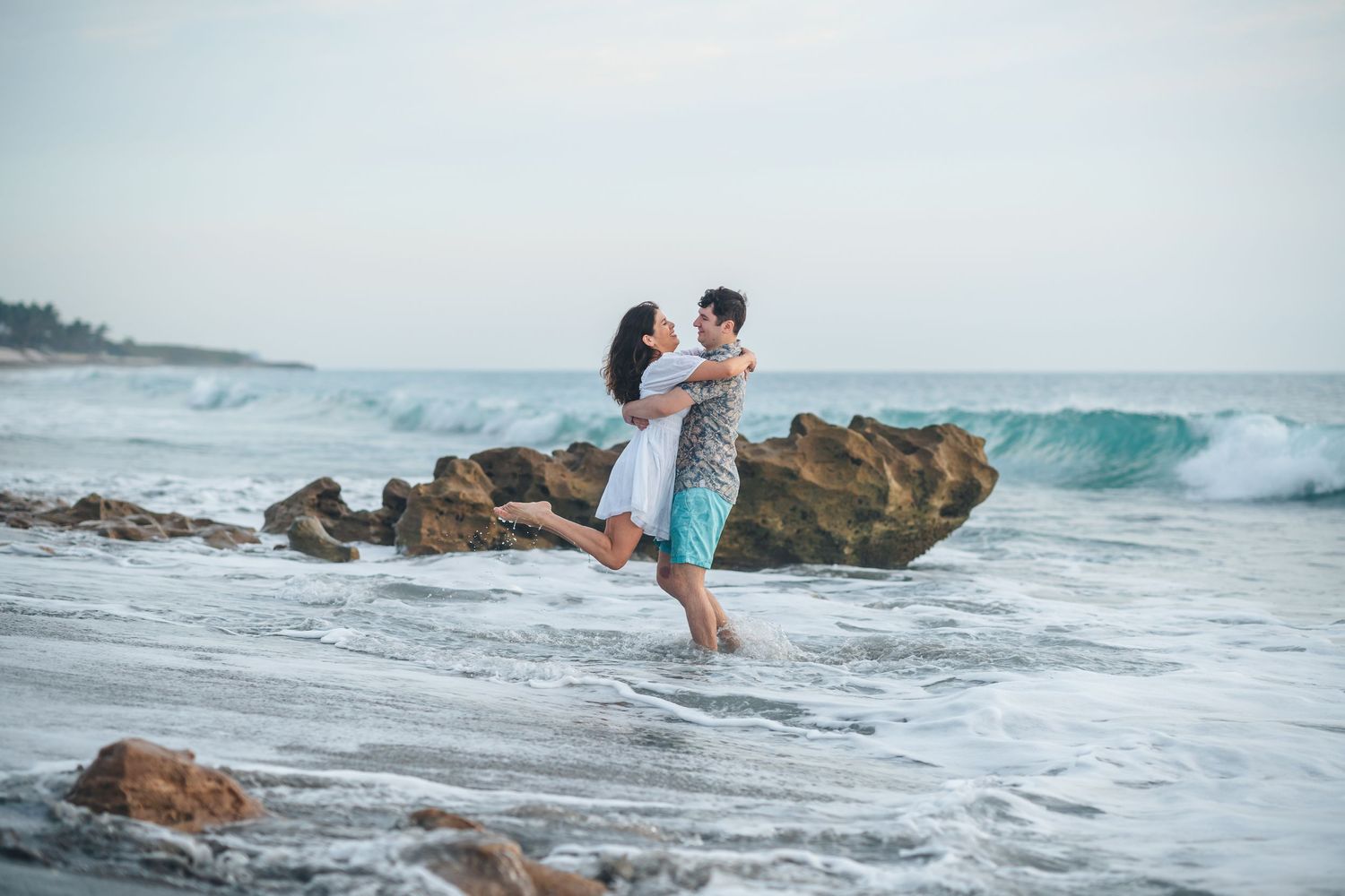 Couple playing and lifting each other on rocky beach with crashing waves.