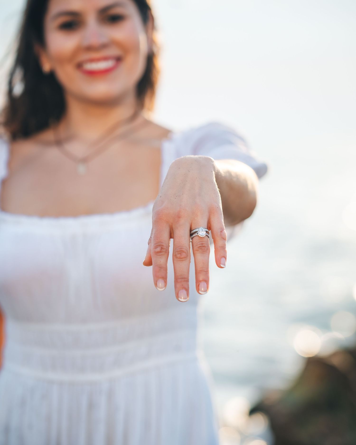 Close up of an engagement ring on a hand with a white dress and ocean background.