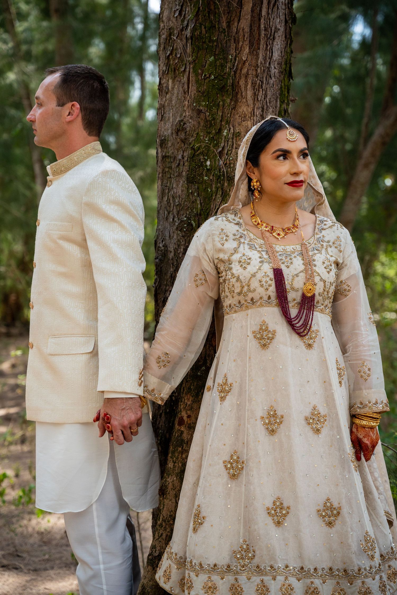 Fusion wedding couple in white and gold attire pose by tree in woodland setting.