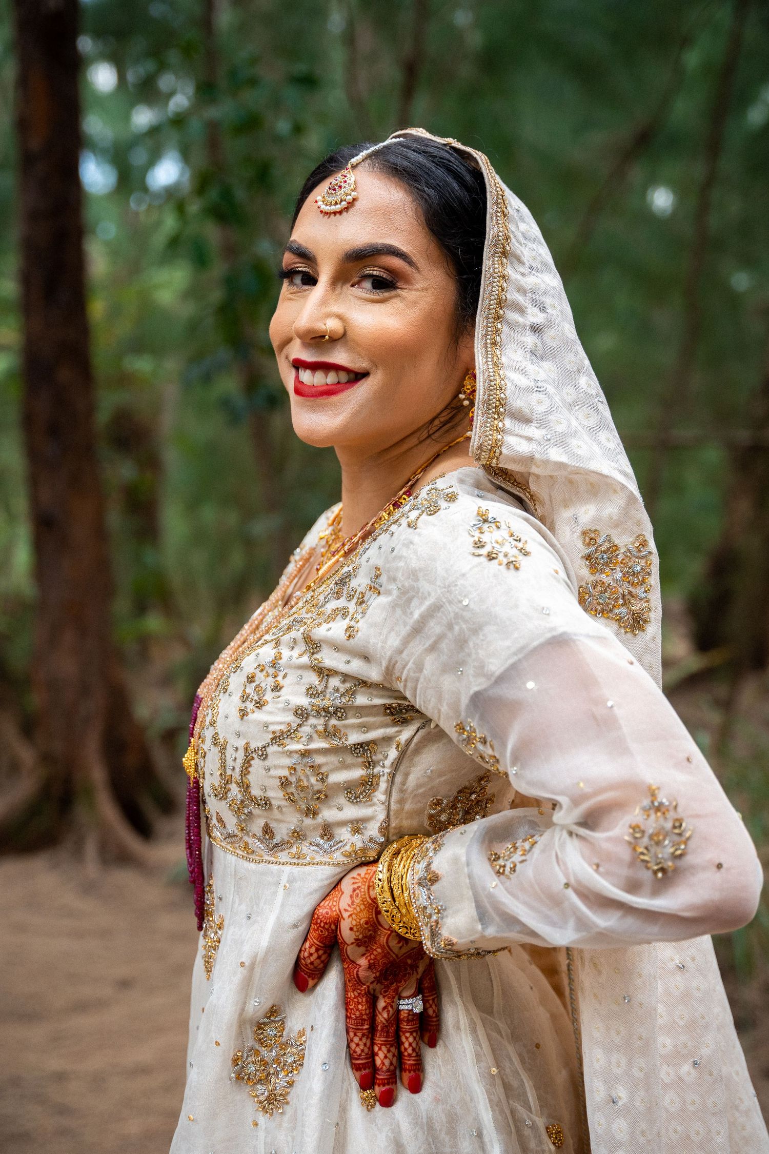 Traditional South Asian bride in white and gold embroidered dress smiles in forest setting.