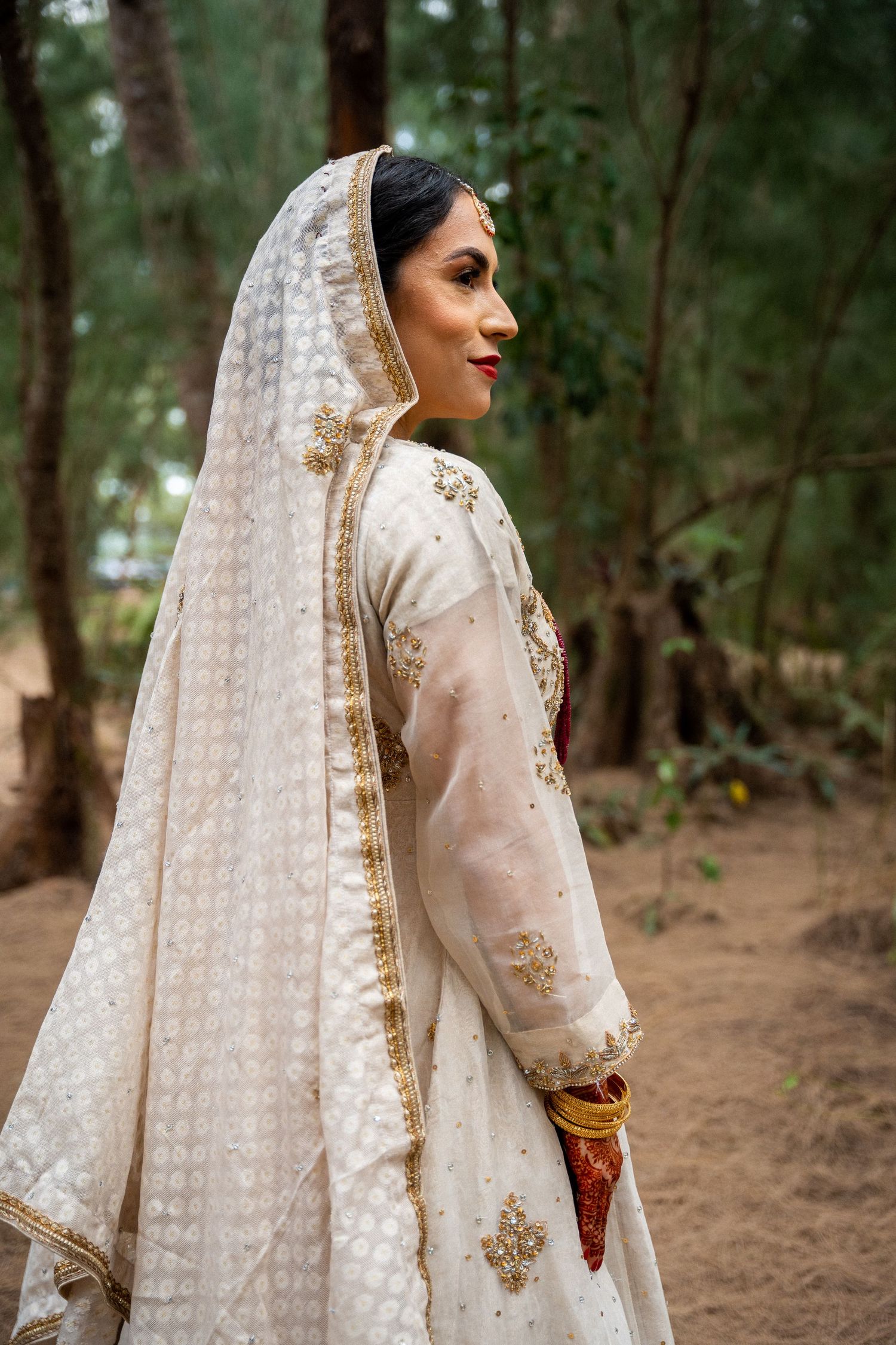 Side profile of bride in ornate white and gold lengha with detailed veil against forest backdrop.