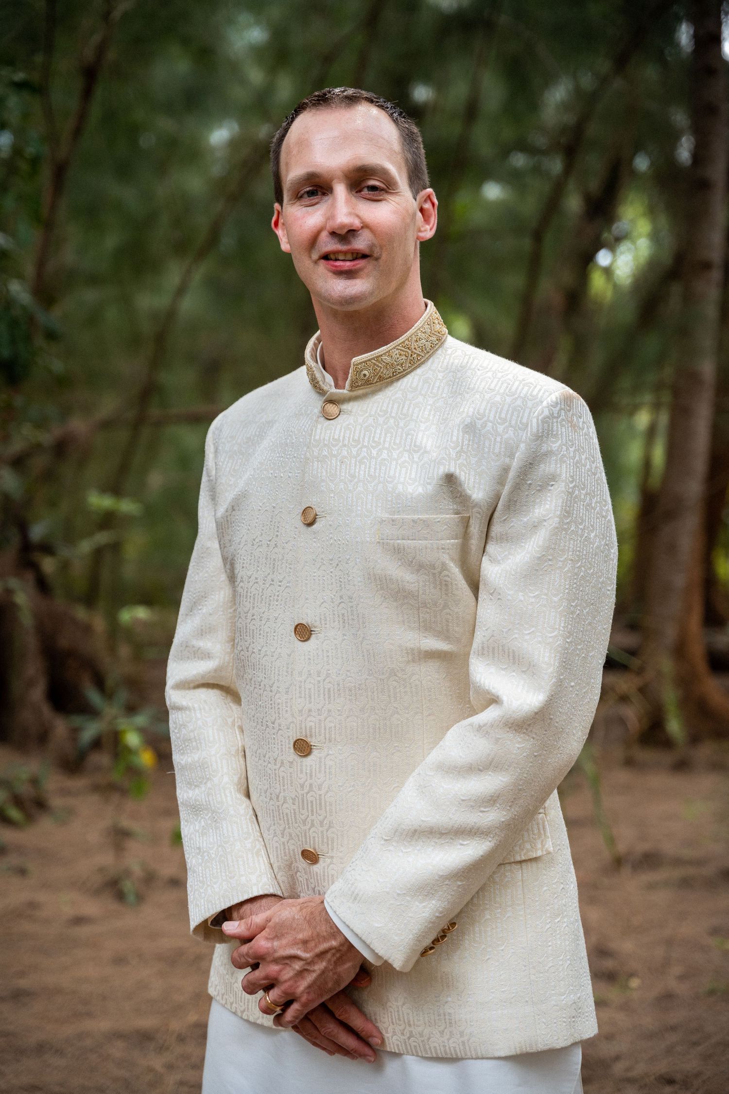 Groom wearing cream Nehru collar jacket and white pants poses in forest setting.