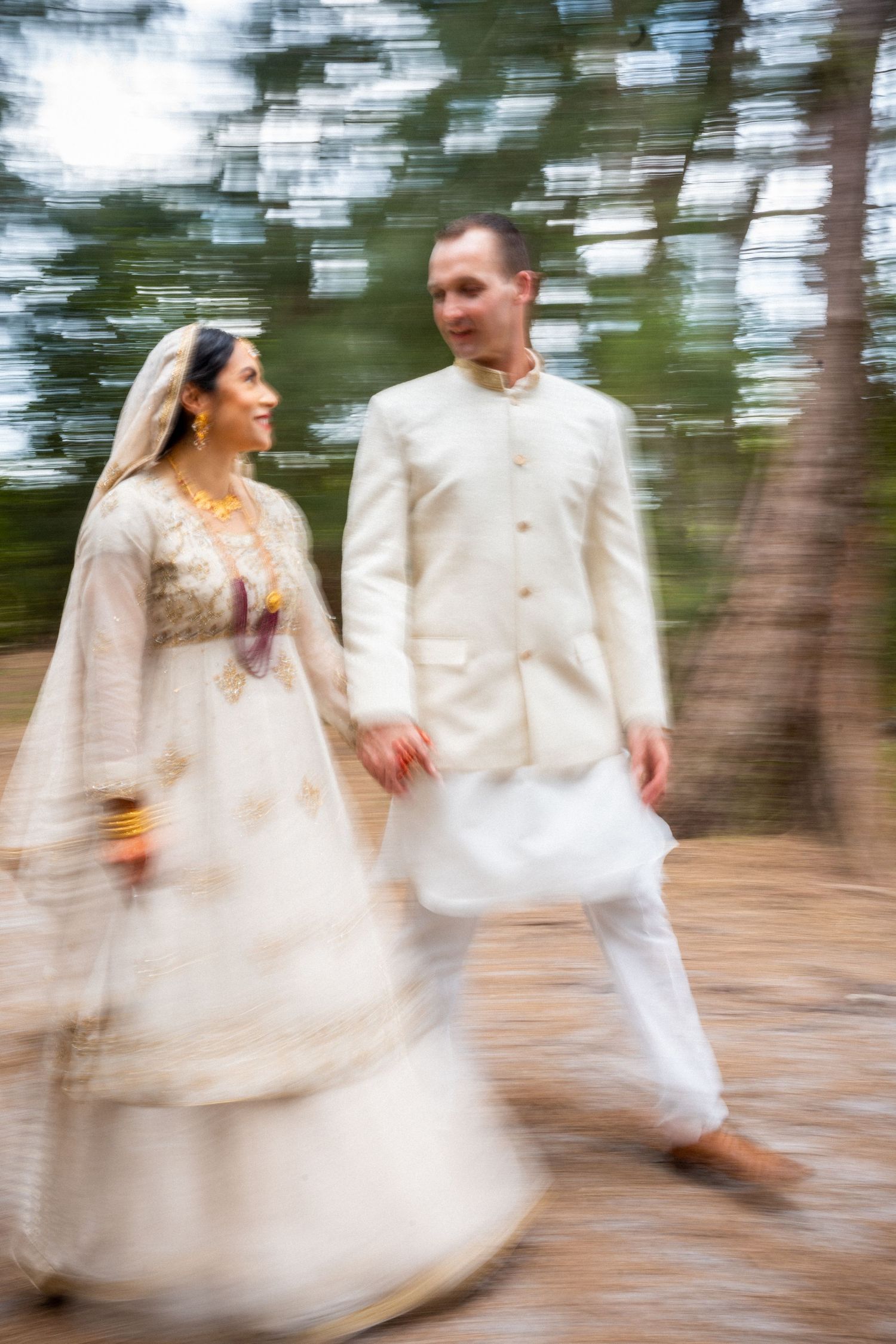 Motion-blur capture of couple in white and gold wedding attire walking through forest.