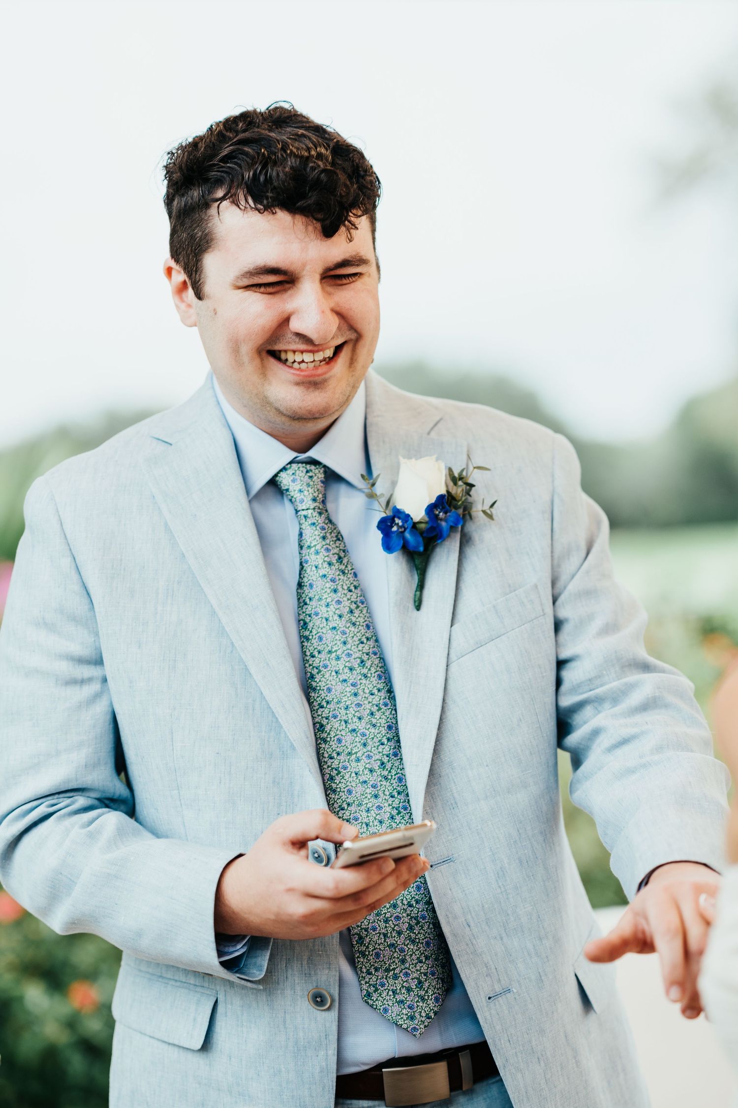 Groom wearing light blue suit and floral tie checking phone during wedding.