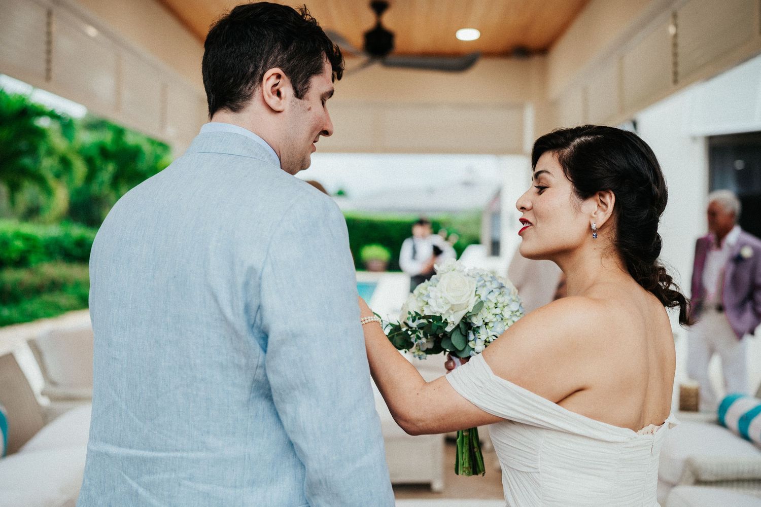 Bride in white dress shares a tender moment with groom in light gray suit while holding a bouquet outdoors.