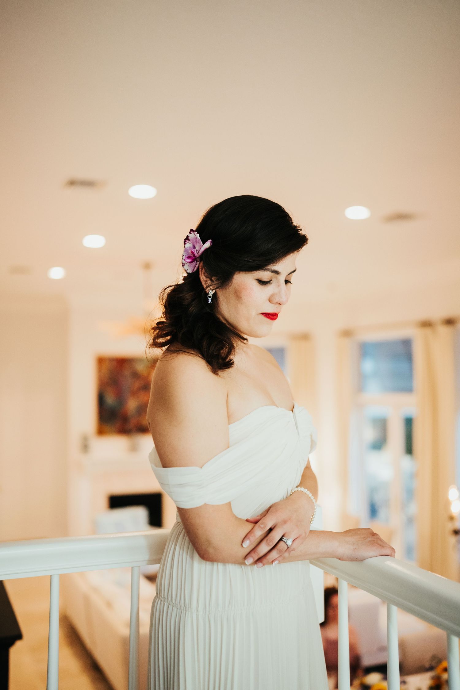 Bride in off-shoulder pleated dress poses elegantly with red lipstick and floral hair accessory.