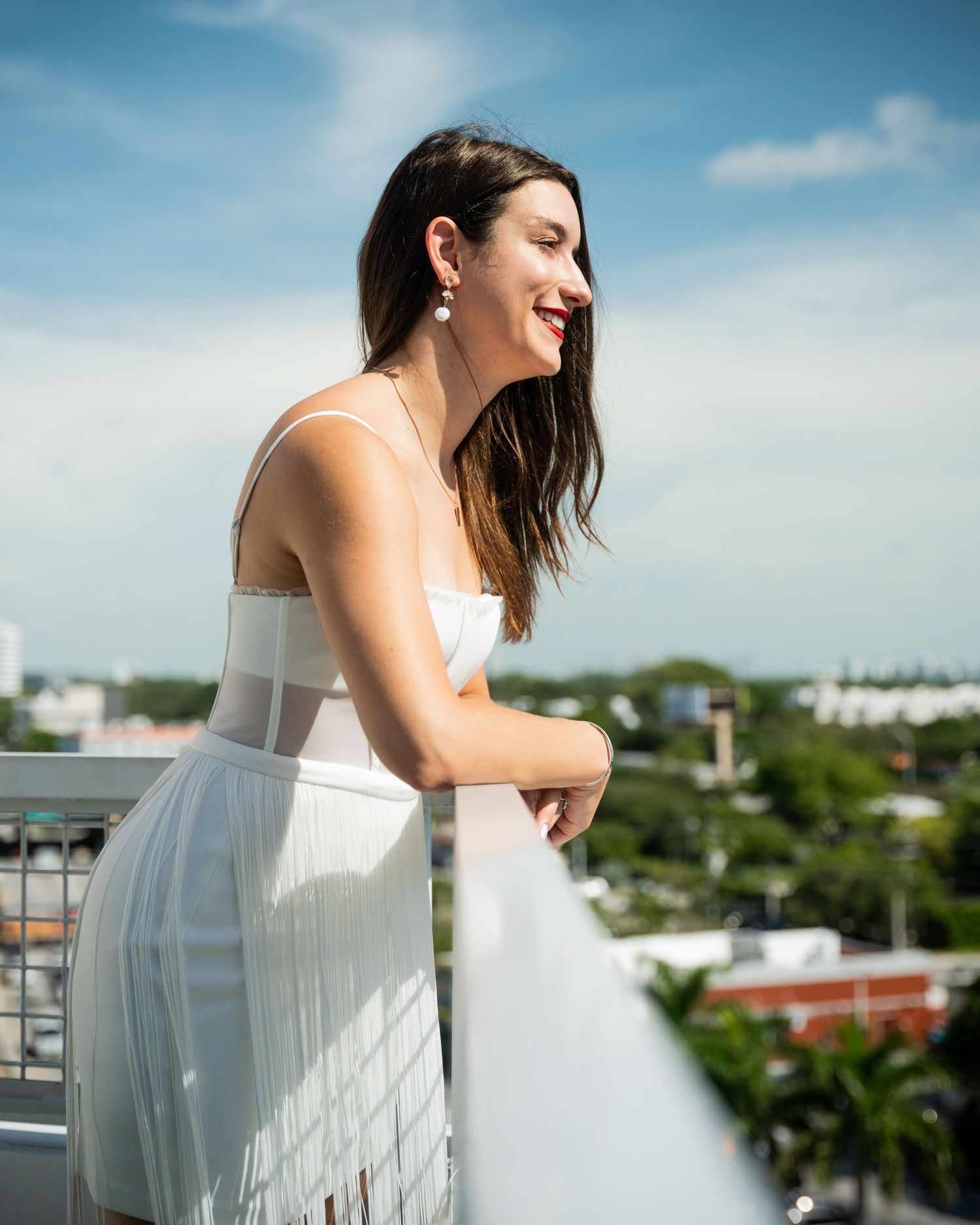 A woman in a white dress smiles while enjoying a sunny day on a balcony.