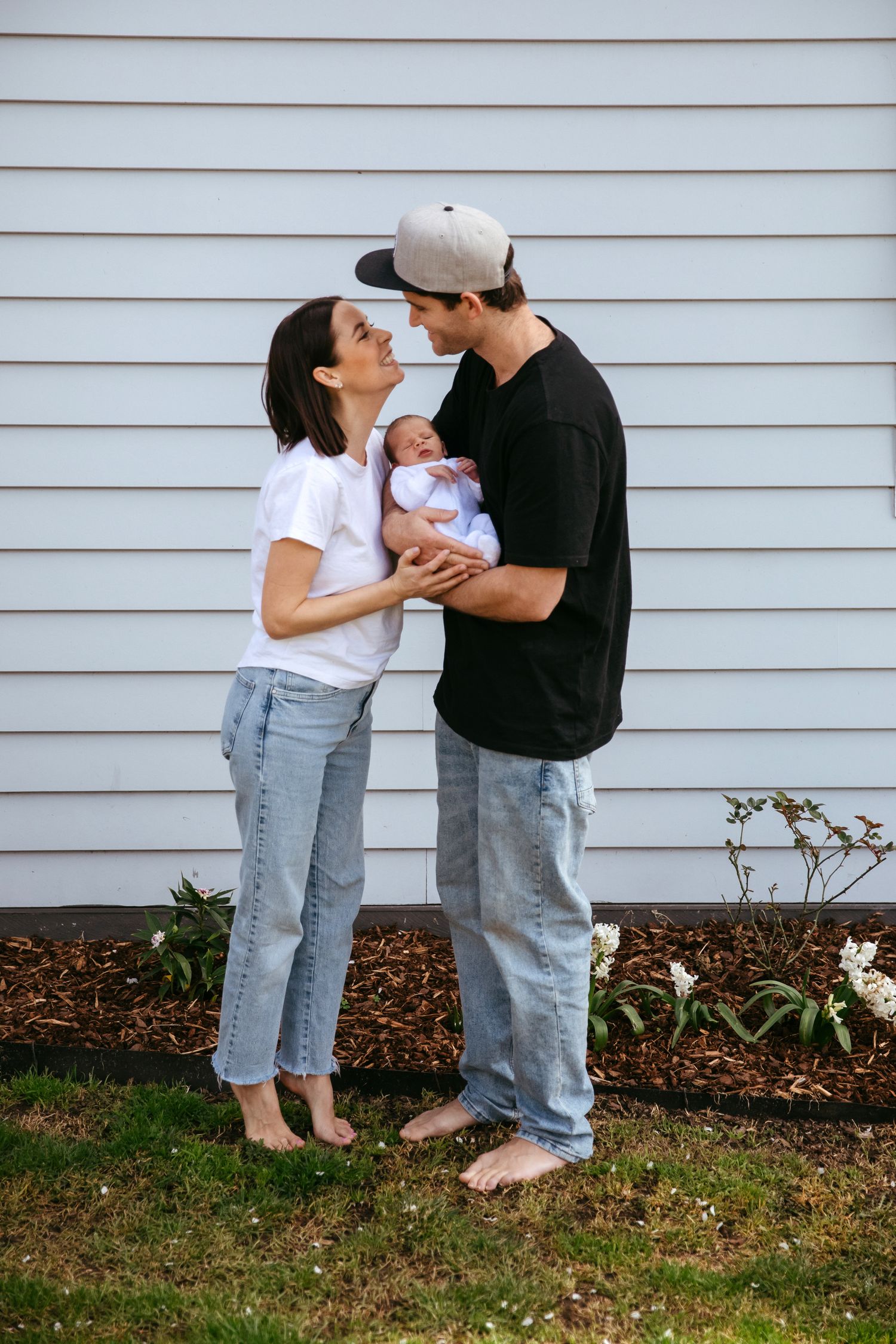 Family shares an intimate moment against white siding of house with flowers in foreground.