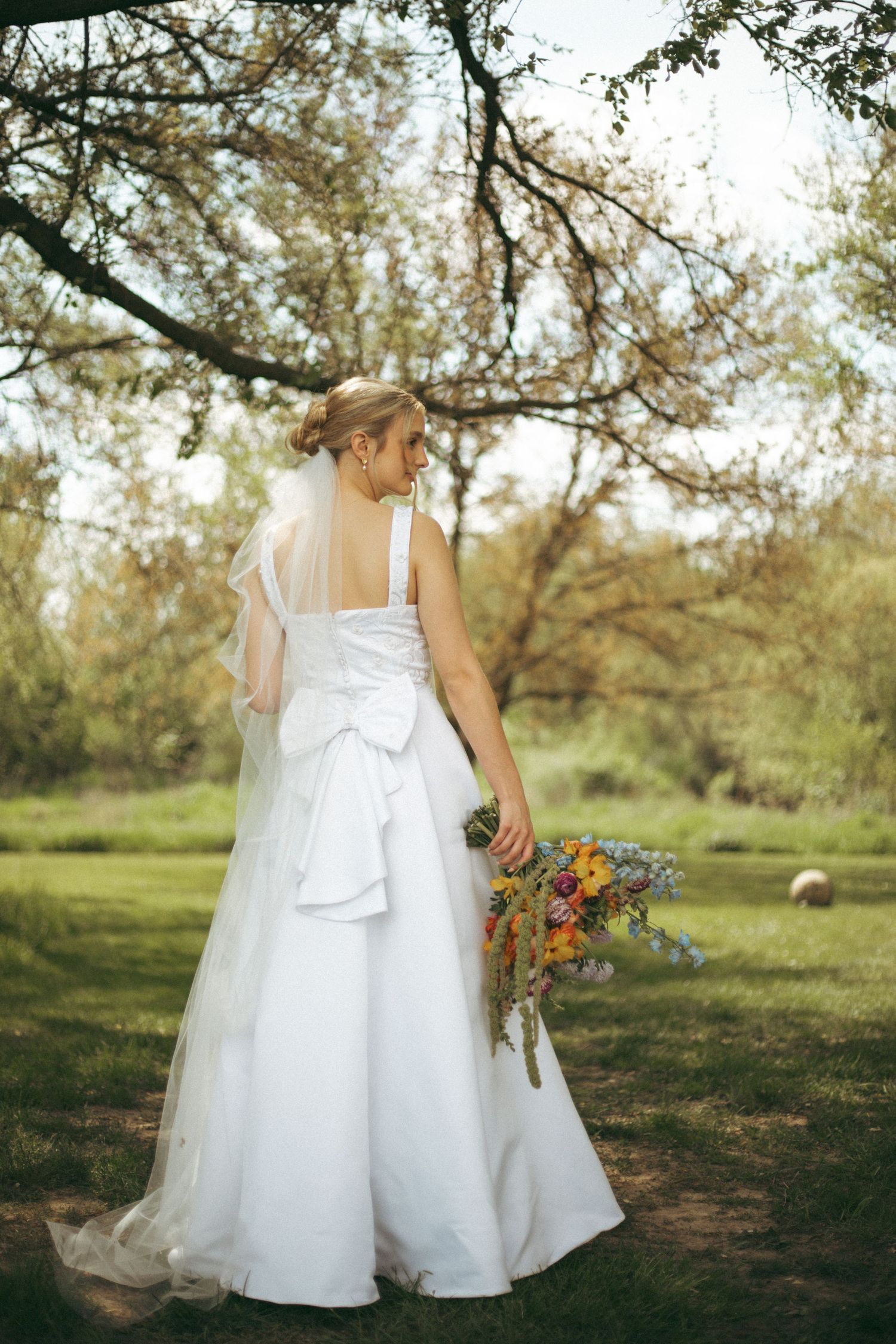 Back view of wedding dress with flowing skirt and bouquet in outdoor garden setting.