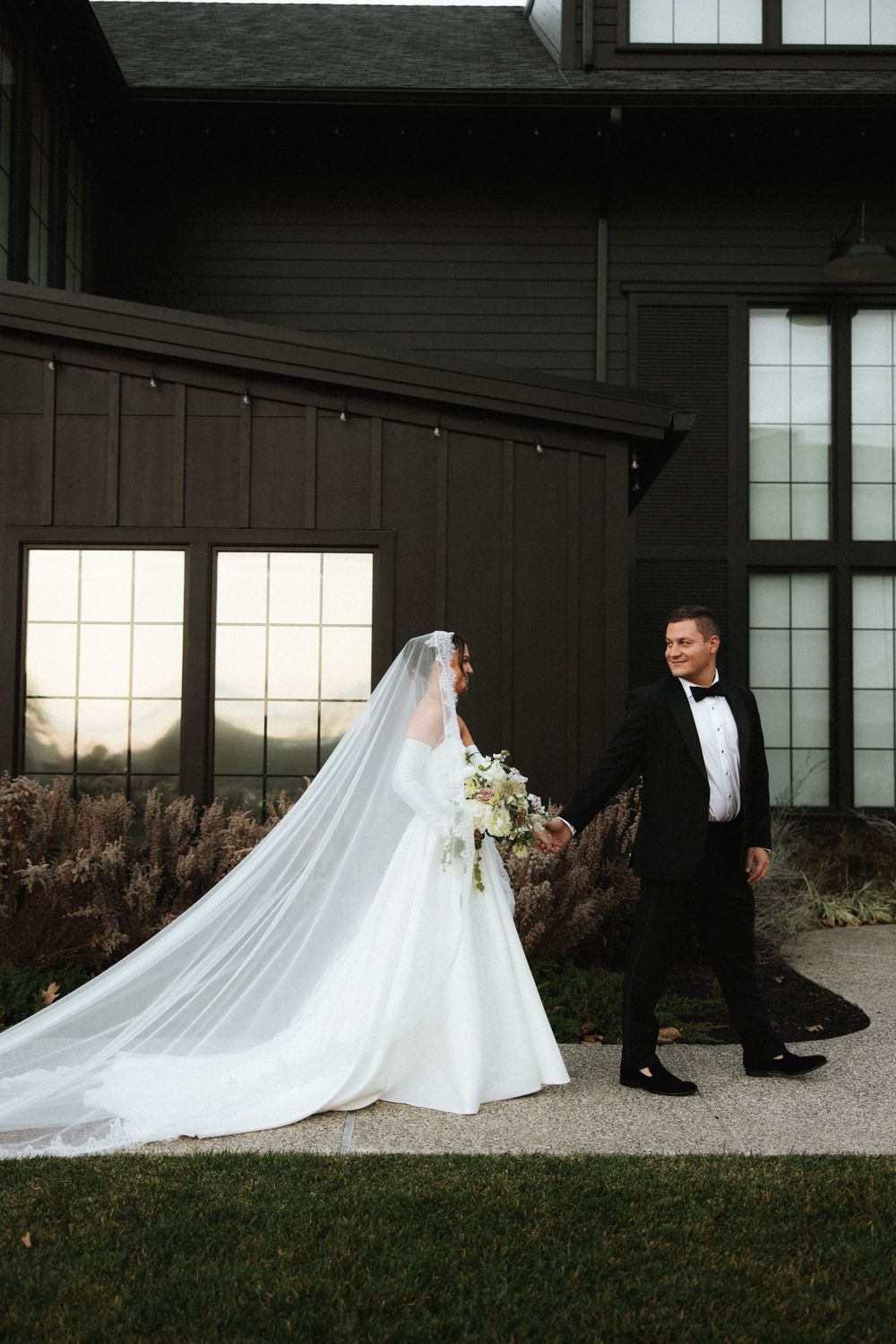 Bride in long veil and groom in tuxedo walking hand in hand outside dark modern barn venue.